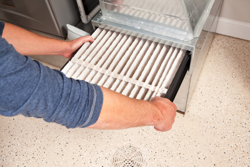 Male hands are installing a furnace air filter. This filter is not brand new but it is relatively clean, it was removed for inspection. This would be a typical filter style found in a high end newer house. The floor is epoxy painted concrete with traction chips and a floor drain.